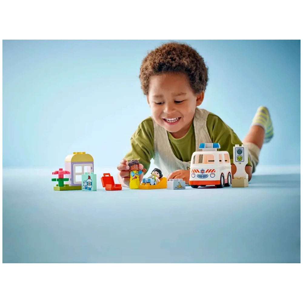 A smiling child with curly hair plays with the LEGO® DUPLO® Ambulance & Driver set, surrounded by colorful blocks, figures, and a toy ambulance on a light blue background.