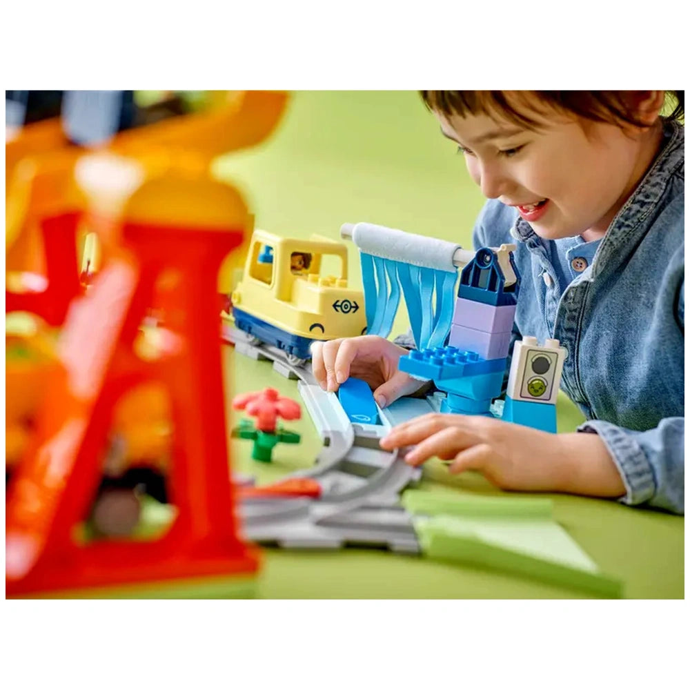A young child smiles while playing with the Lego Duplo Big Interactive Community Train set, assembling train tracks with colorful blocks and figures.