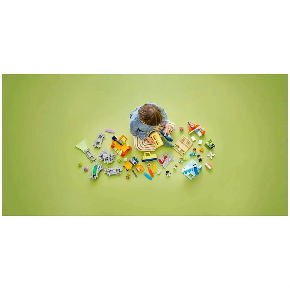 A young child sits on a green surface, engaged with the Lego Duplo Big Interactive Community Train set, surrounded by colorful building elements, figures, and a train station, fostering imaginative play and creativity.