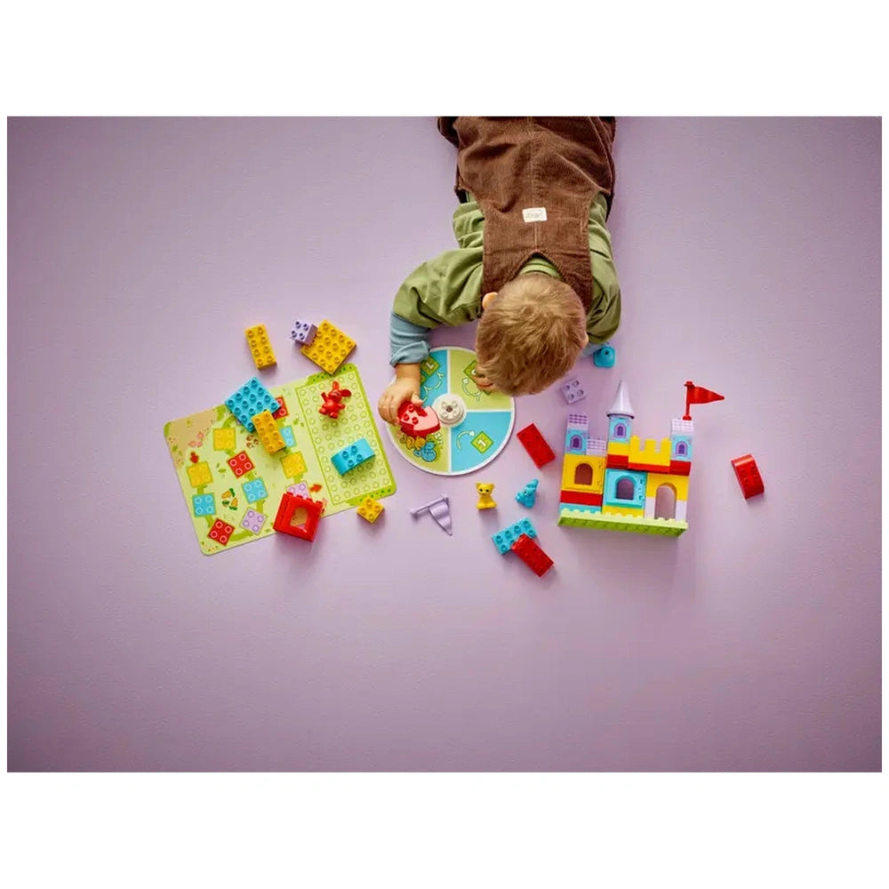 A child lays on a purple surface, engaging with the LEGO® DUPLO® Hopsy’s Castle Game. Colorful building blocks, a game board, and animal figures are scattered around, highlighting fun, creative play.
