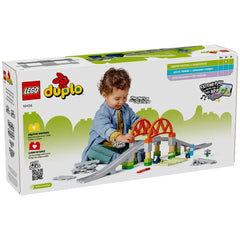 A toddler in a blue shirt and denim jacket plays with the LEGO® DUPLO® Train Bridge and Tracks Expansion Set, featuring a raised bridge, track pieces, and colorful blocks. The child engages creatively, enhancing motor skills and imagination in a vibrant play scene.