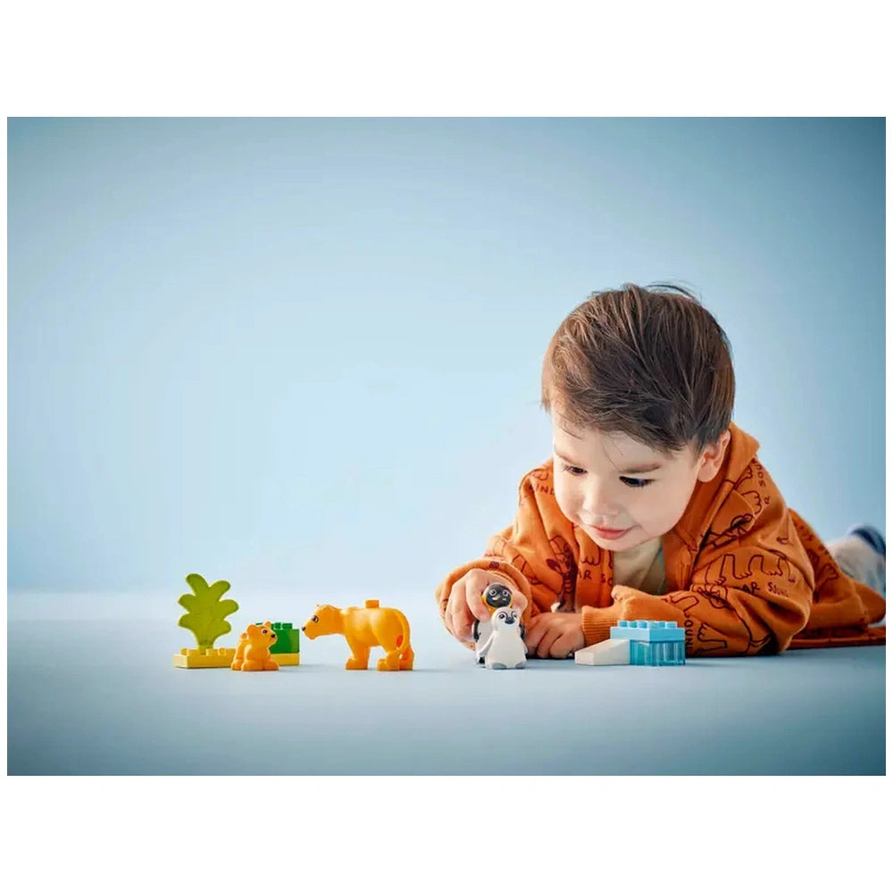 A young child in an orange jacket plays on a blue surface with colorful LEGO® DUPLO® animal figures, including penguins and lions, alongside a green tree and building blocks.