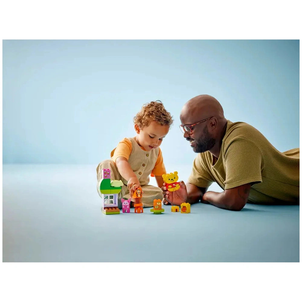 A toddler and an adult man play with the LEGO® DUPLO® Winnie the Pooh’s Birthday Party set on a light blue floor. The toddler, wearing an orange shirt, interacts with colorful building blocks, including characters like Winnie the Pooh, Tigger, and Piglet. The man smiles, encouraging the child's imaginative play, surrounded by vibrant toy elements that inspire creativity and storytelling.