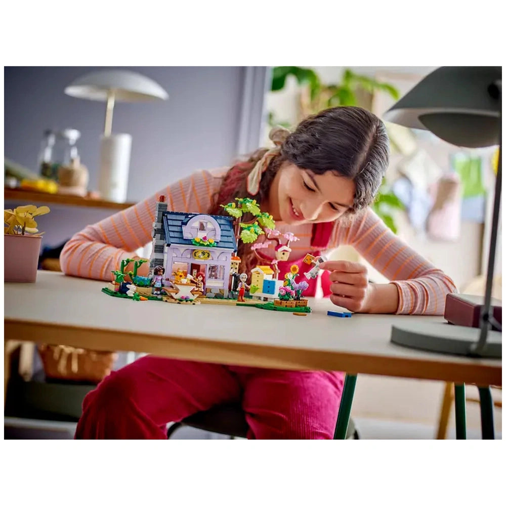 A young girl with long, curly hair smiles while building the LEGO Friends Beekeepers' House and Flower Garden set, featuring colorful flowers and a two-story house on a table.