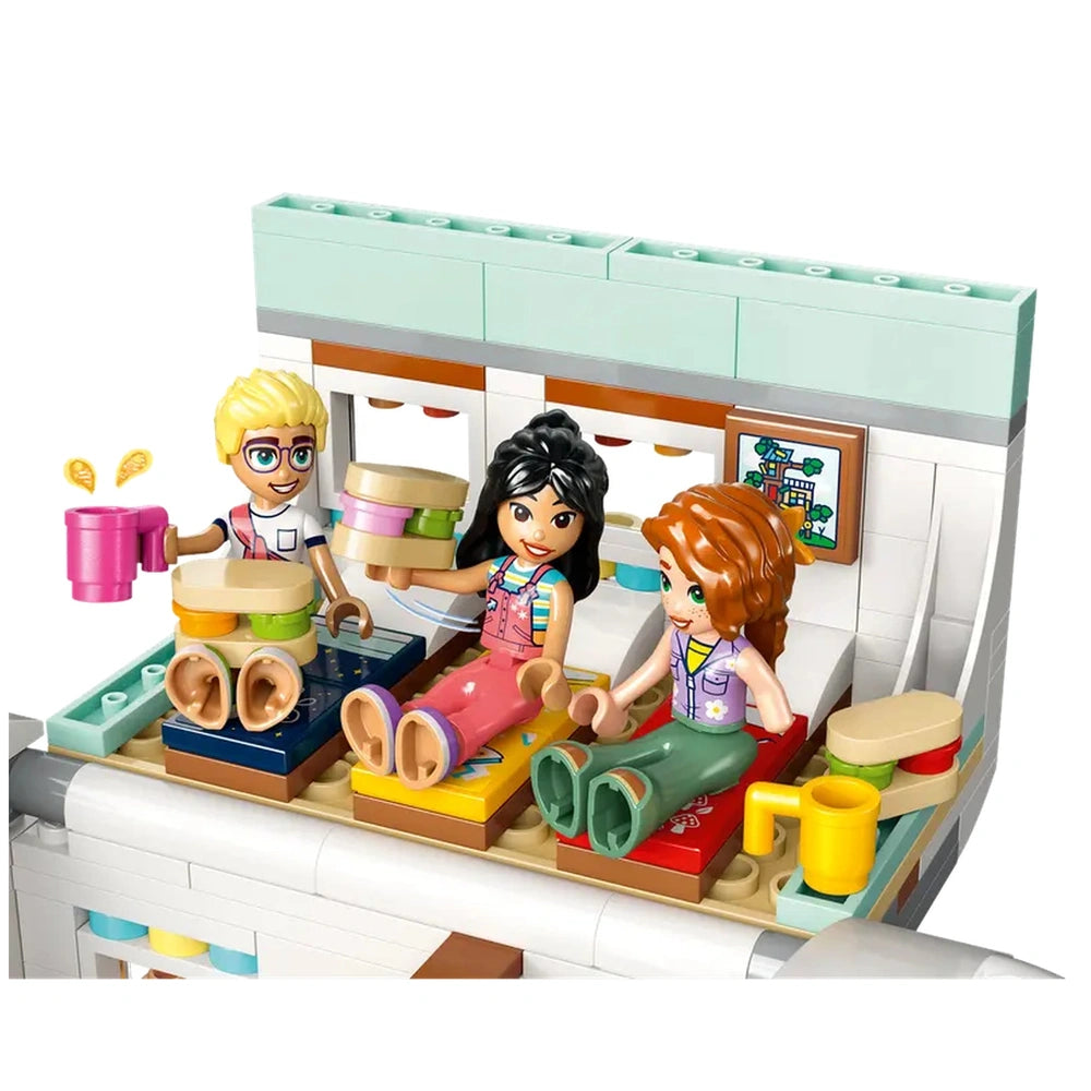 Three LEGO minifigures enjoy a cozy indoor scene in the camper van, sitting on colorful beds with playful accessories. The girl on the left holds a pink cup while a boy lifts a tray. The vibrant interior features window details and a fun decorative picture on the wall, creating an inviting atmosphere for imaginative play.