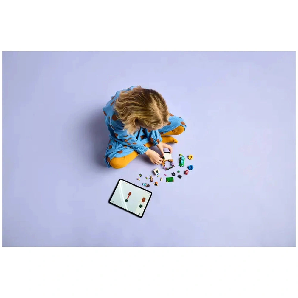 A child sits cross-legged on a lavender floor, engaged with LEGO pieces scattered around. An iPad displays colorful building instructions, while the child carefully assembles a toy.