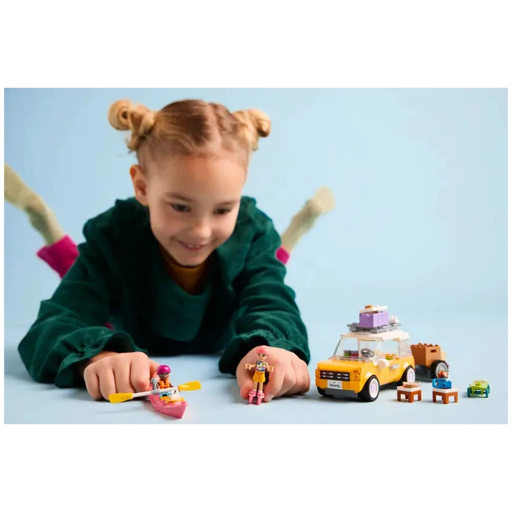 A child with two playful hairstyles is lying on a light blue surface, smiling as they engage with a LEGO Friends travel car and minidolls. The scene includes a pink kayak and other colorful accessories, showcasing a vibrant, imaginative play setup.