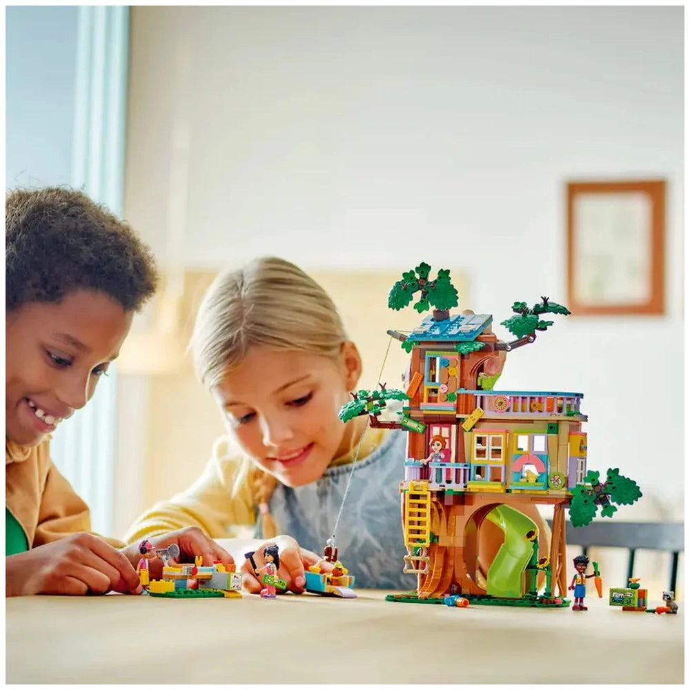 Two children, a boy and a girl, excitedly play with the LEGO Friends Friendship Tree House Hangout set, featuring a colorful treehouse with green leaves, a slide, and various figures. The boy, with curly hair, is focused on a small build, while the girl, with straight hair, smiles at her creation on the table.