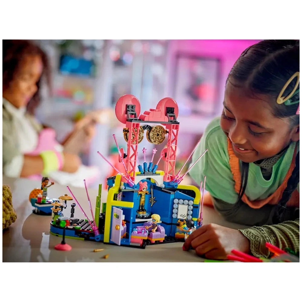 A young girl smiles as she interacts with the colorful LEGO® Friends Heartlake City Music Talent Show set, featuring a vibrant stage, mini-dolls, and musical accessories. In the background, another child plays a pink guitar, adding to the lively atmosphere.