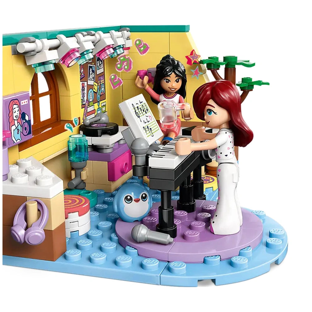 Two LEGO Friends minidolls, one with dark hair and the other with red hair, engage in a playful music session at a colorful room. The scene includes a piano, a cute blue bunny figure, and vibrant music-themed decor on the walls, highlighting creativity and friendship.