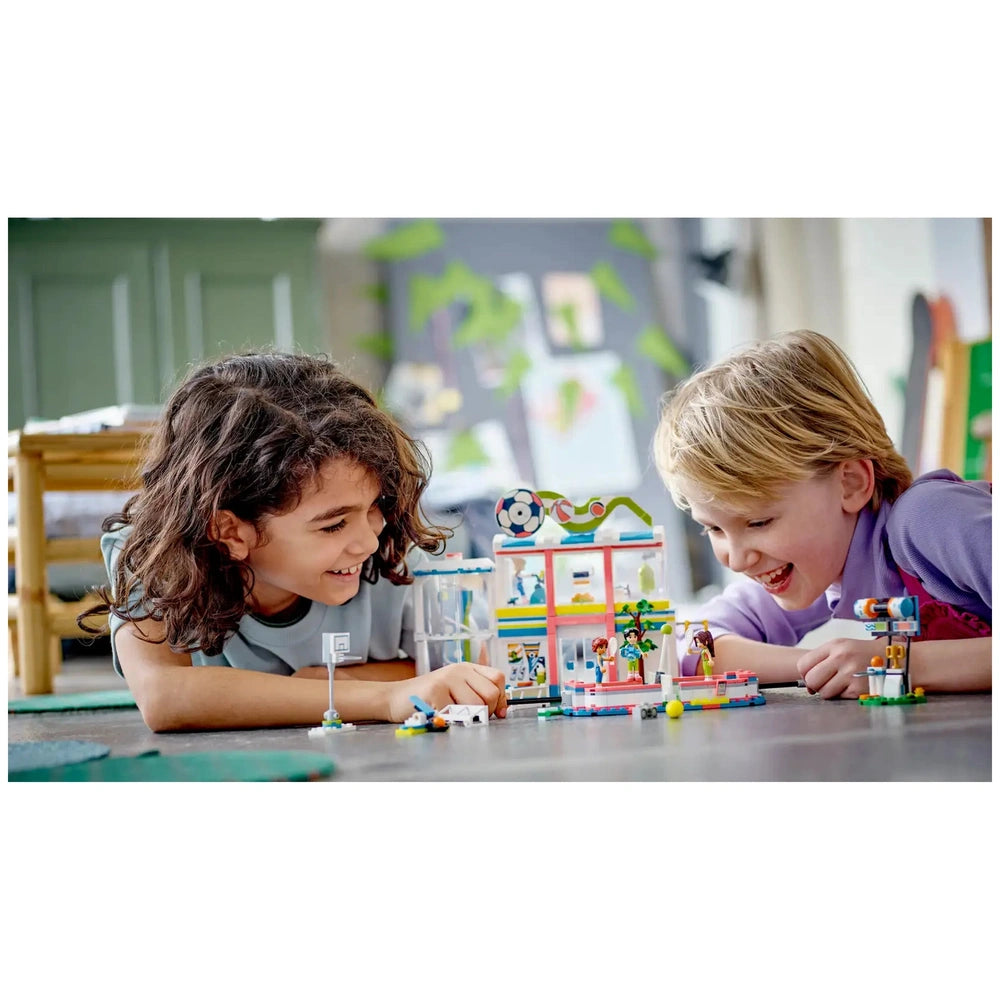 Two children play together on a colorful mat, engaged with the LEGO® Friends Sports Center set, featuring mini-dolls and various sports accessories, fostering creativity and teamwork.