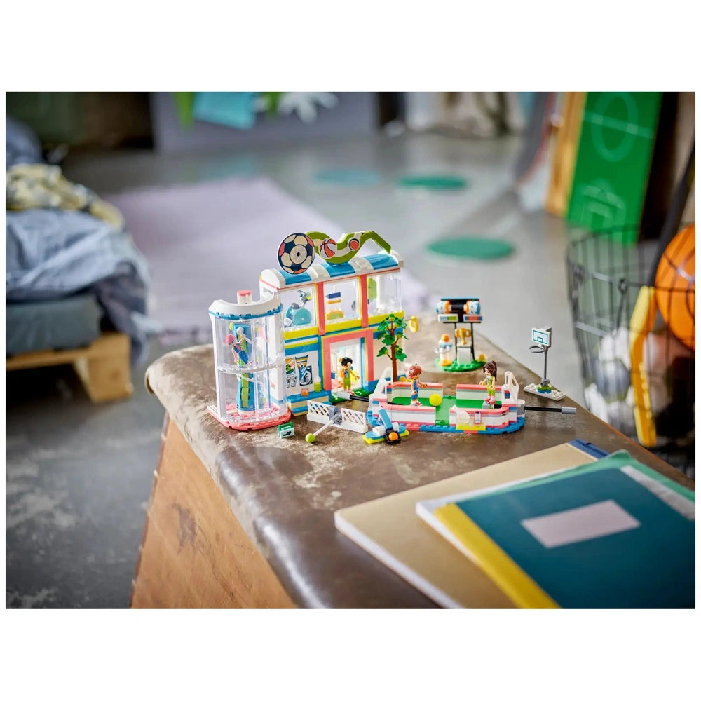 Colorful LEGO® Friends Sports Center set on a wooden table, featuring a variety of sports scenes with mini-dolls playing soccer and basketball, surrounded by accessories like balls and nets.