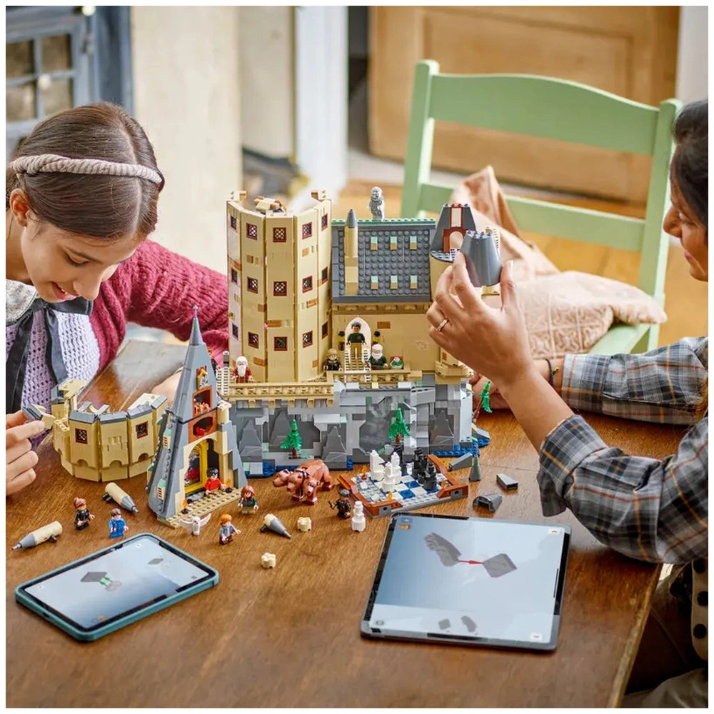 Two children assembling the LEGO Harry Potter Hogwarts Castle set on a wooden table, with detailed minifigures and colorful bricks scattered around. Two tablets displaying building instructions are visible.