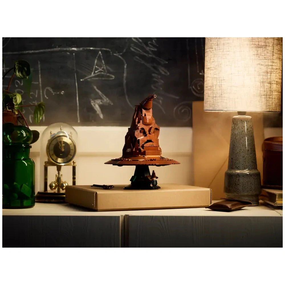 LEGO Harry Potter Talking Sorting Hat construction set displayed on a table, surrounded by a green plant, clock, and a lamp, with a chalkboard in the background.