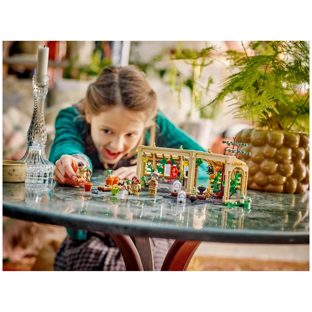 A child is excitedly playing with the LEGO Hogwarts™ Castle: Herbology Class set on a glass table, featuring detailed miniatures of plants and figures in a vibrant greenhouse.