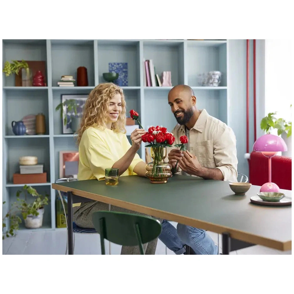 A woman in a yellow shirt and a man in a beige shirt joyfully arrange a vibrant bouquet of red LEGO® roses at a modern green table. A glass and small dish are nearby, while decorative items fill the shelves behind them.