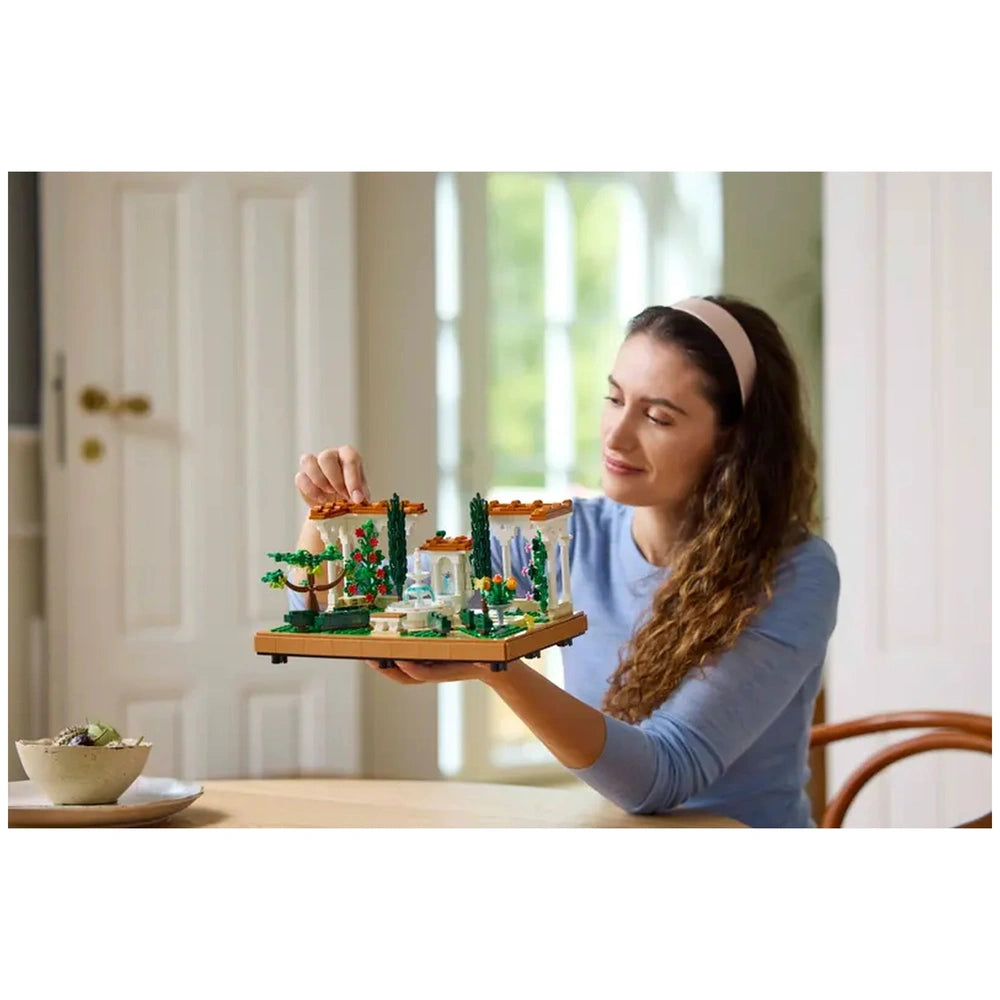 A woman with curly hair is gently adjusting a LEGO® Icons Fountain Garden construction set, showcasing intricate landscaping and vibrant flora. She smiles as she holds the display, which features a tiered fountain, decorative pathways, and charming pergolas. A bowl with greenery is placed on the table in front of her, with soft natural light filtering through a nearby window.