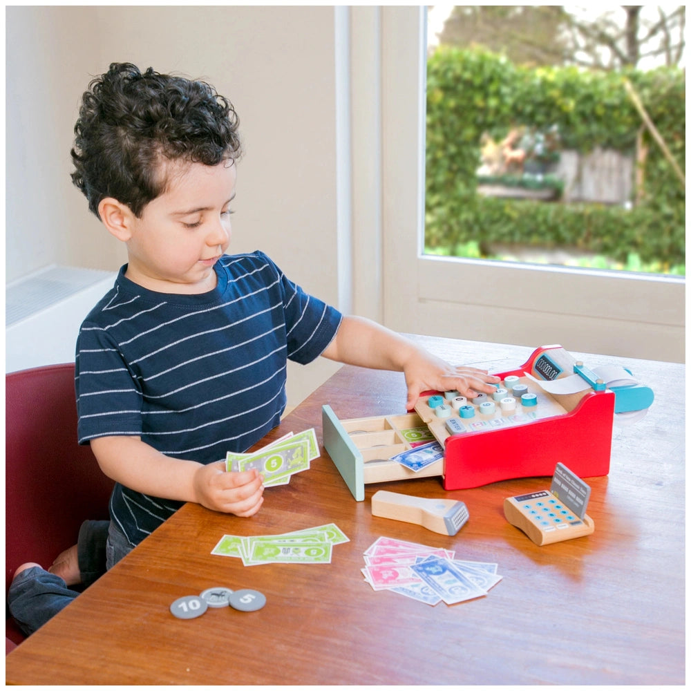 Red and blue cash register toy on a wooden table with paper money, coins, and a child playing joyfully.