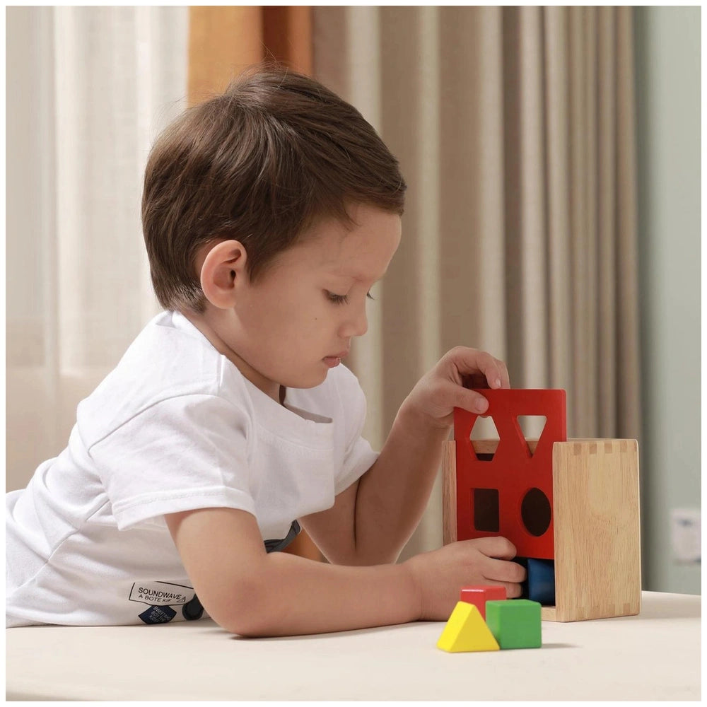 Colorful shape sorter toy with red, blue, and yellow blocks on a wooden table, played with by a child in a white shirt.