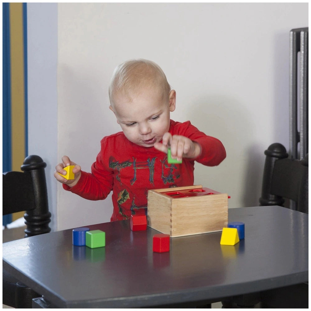 Colorful shape sorter toy on a wooden table with a young child playing, surrounded by plastic cubes and a metal chair.