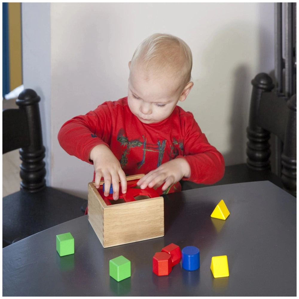 Colorful shape sorter by New Classic Toys with red, yellow, and green pieces on a brown wooden box beside a young child.