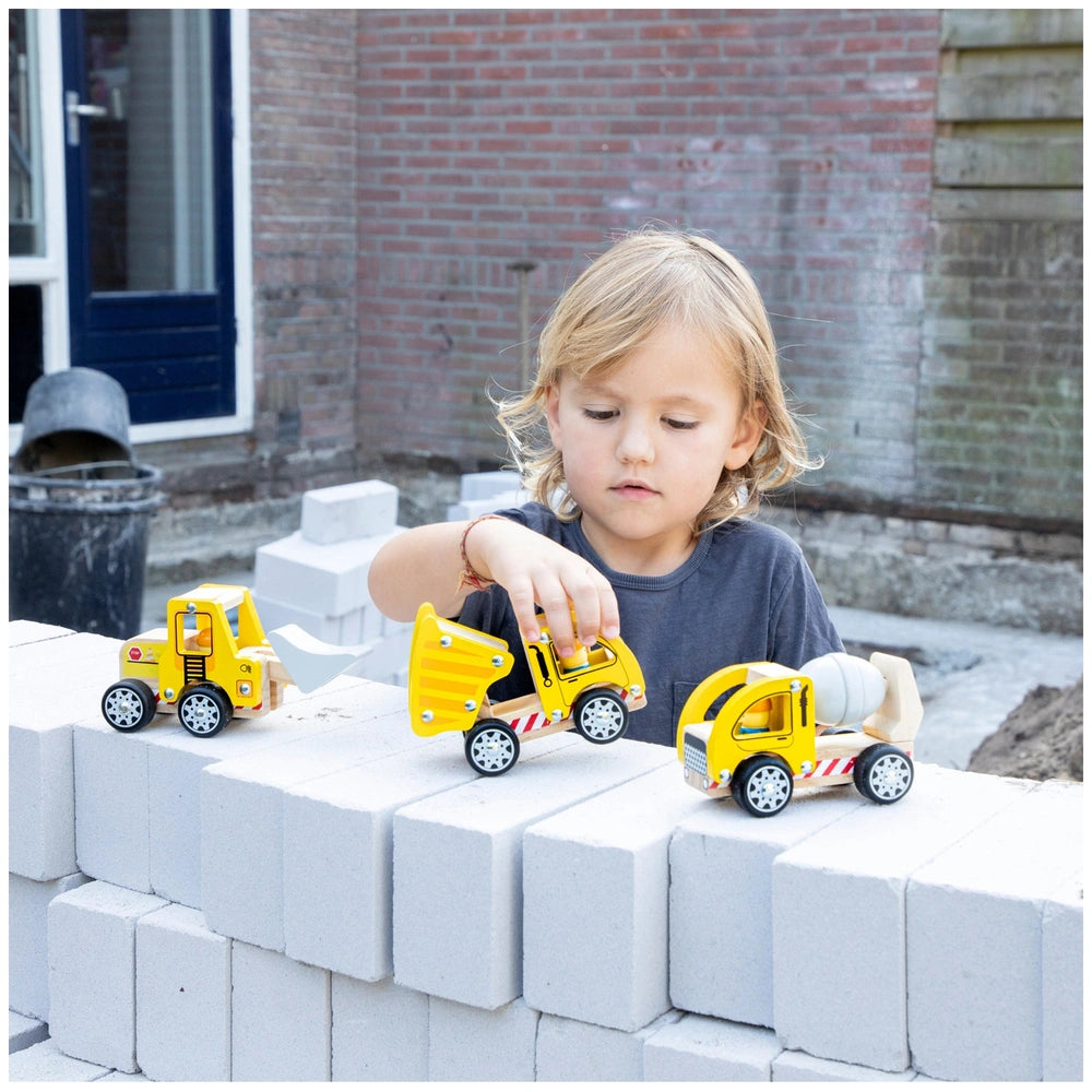 Child with blonde hair plays with yellow and red toy construction vehicles on a concrete ground against a brick wall.