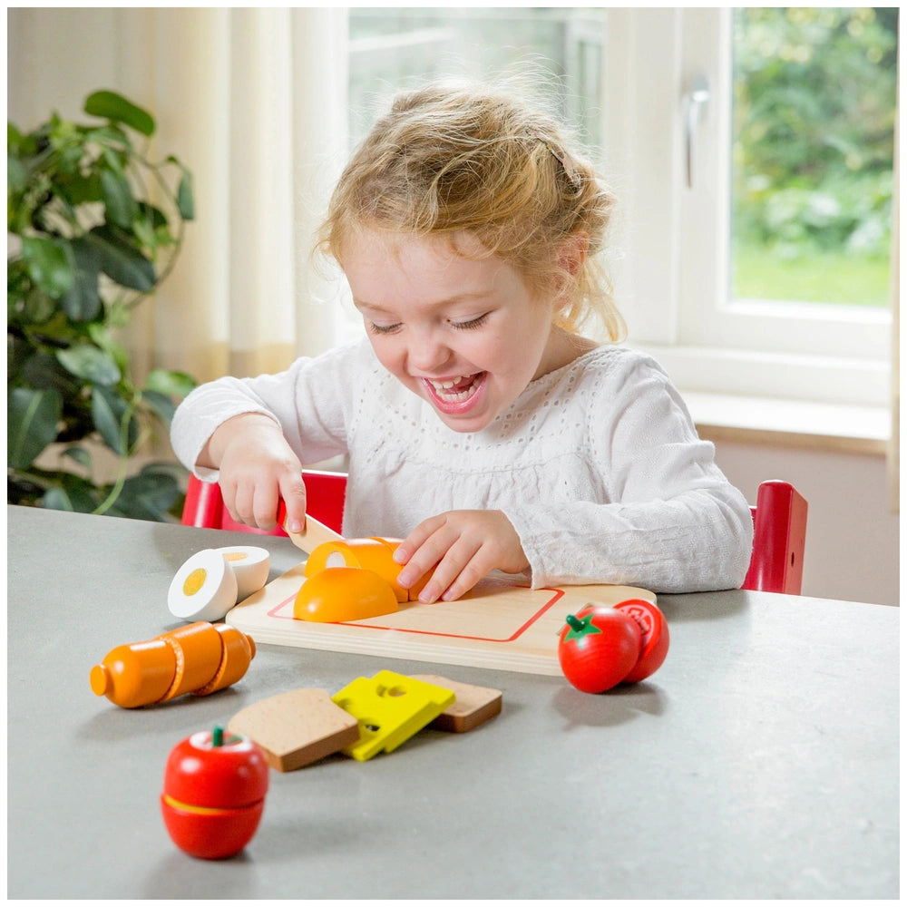Young girl playing with New Classic Toys wooden cutting board, yellow plastic knife, and colorful pretend food items.