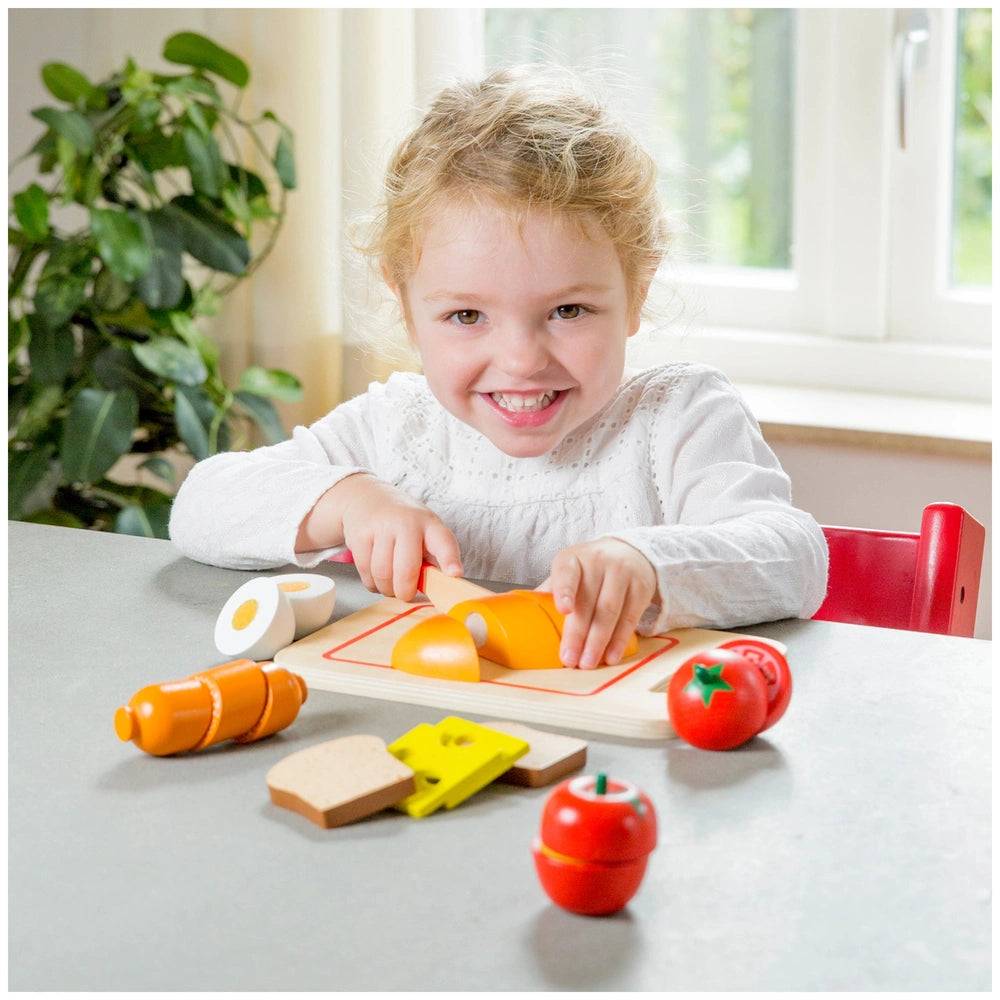 New Classic Toys breakfast cutting set with colorful plastic food items displayed on a wooden table, featuring a young girl p