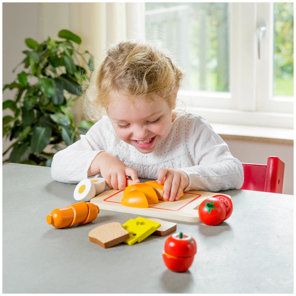 New Classic Toys Cuttingset features colorful plastic food items on a wooden table, with children engaged in pretend play.