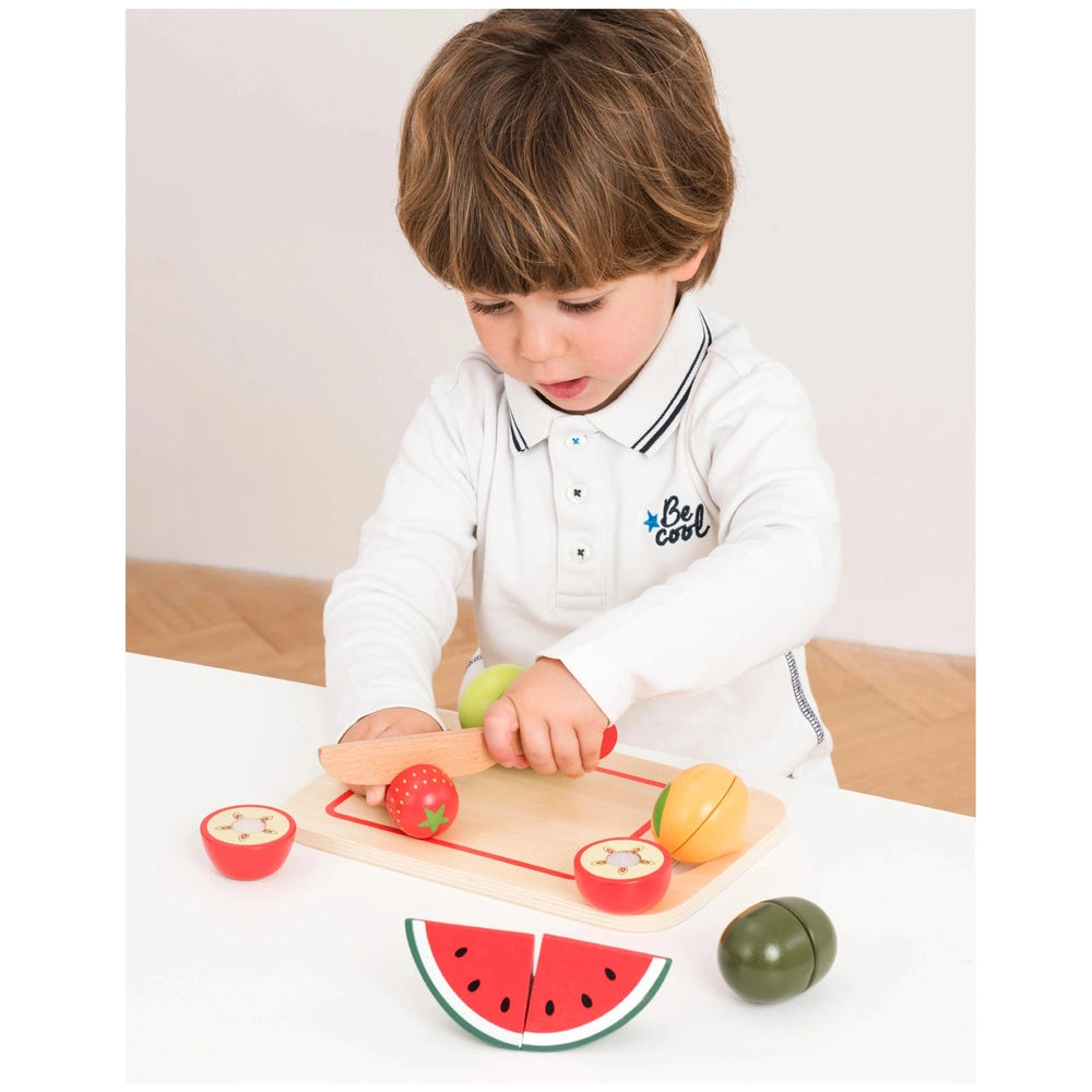 New Classic Toys fruit cutting set with wooden board, plastic knife, and colorful fruits in a playful scene with a child.