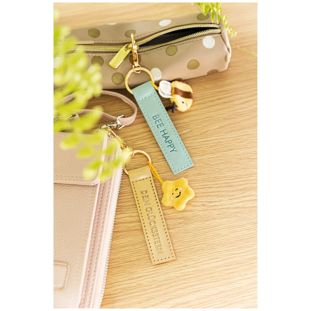 Bee Happy keyholder attached to a beige purse with a gold zipper, featuring a yellow star charm and blue tag, beside a green 