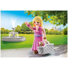 Playmobil It-Girl doll in a pink dress holds a white basket with a small brown dog, set against a green lawn and blue sky.