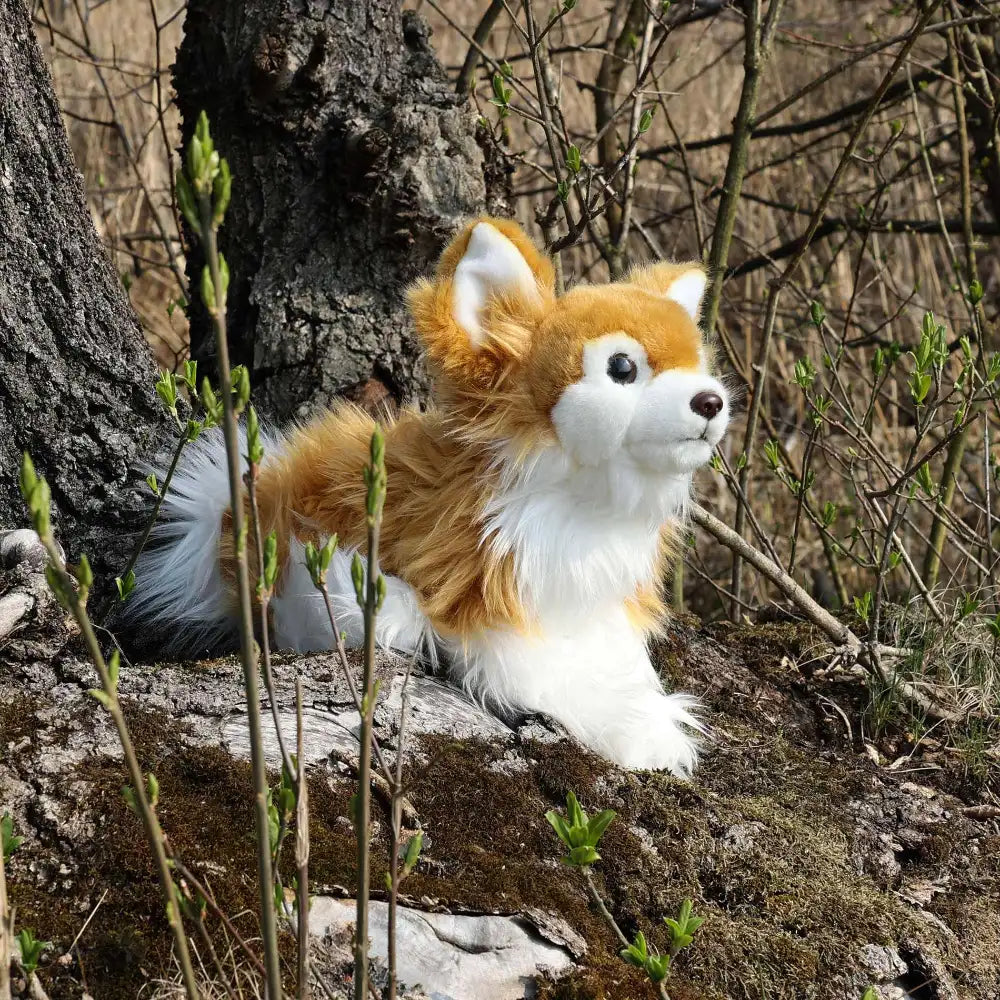 Plush fox toy with orange and white fur sits on moss-covered rock in a natural outdoor setting with green plants and a tree.