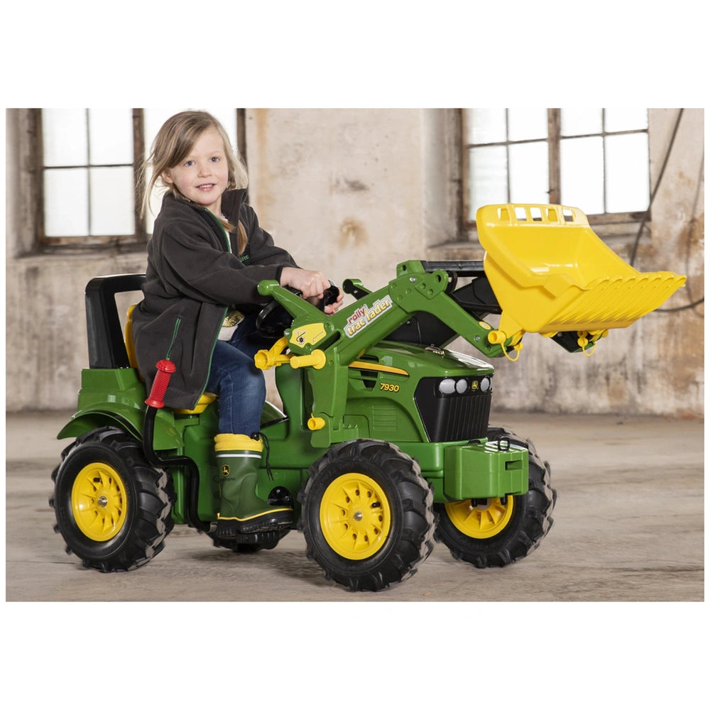 A young girl smiles while sitting on a green and yellow toy tractor with a bucket, indoors on a concrete floor.