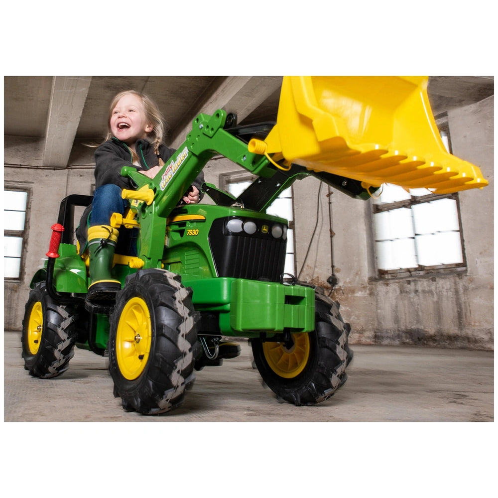 Green and yellow toy tractor with a smiling girl in a jacket and boots sitting on it indoors.