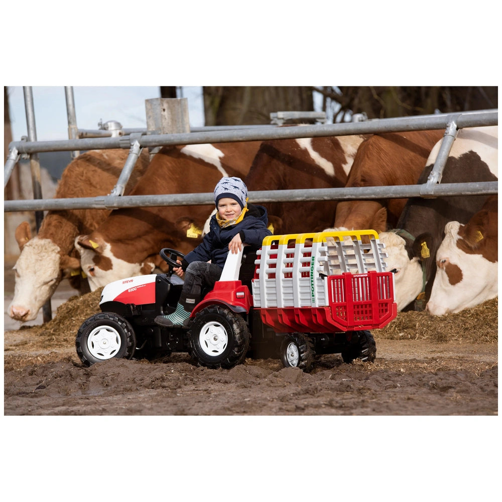A child in a winter hat rides a red and white toy tractor with a basket, surrounded by brown and white cows on a muddy farm.
