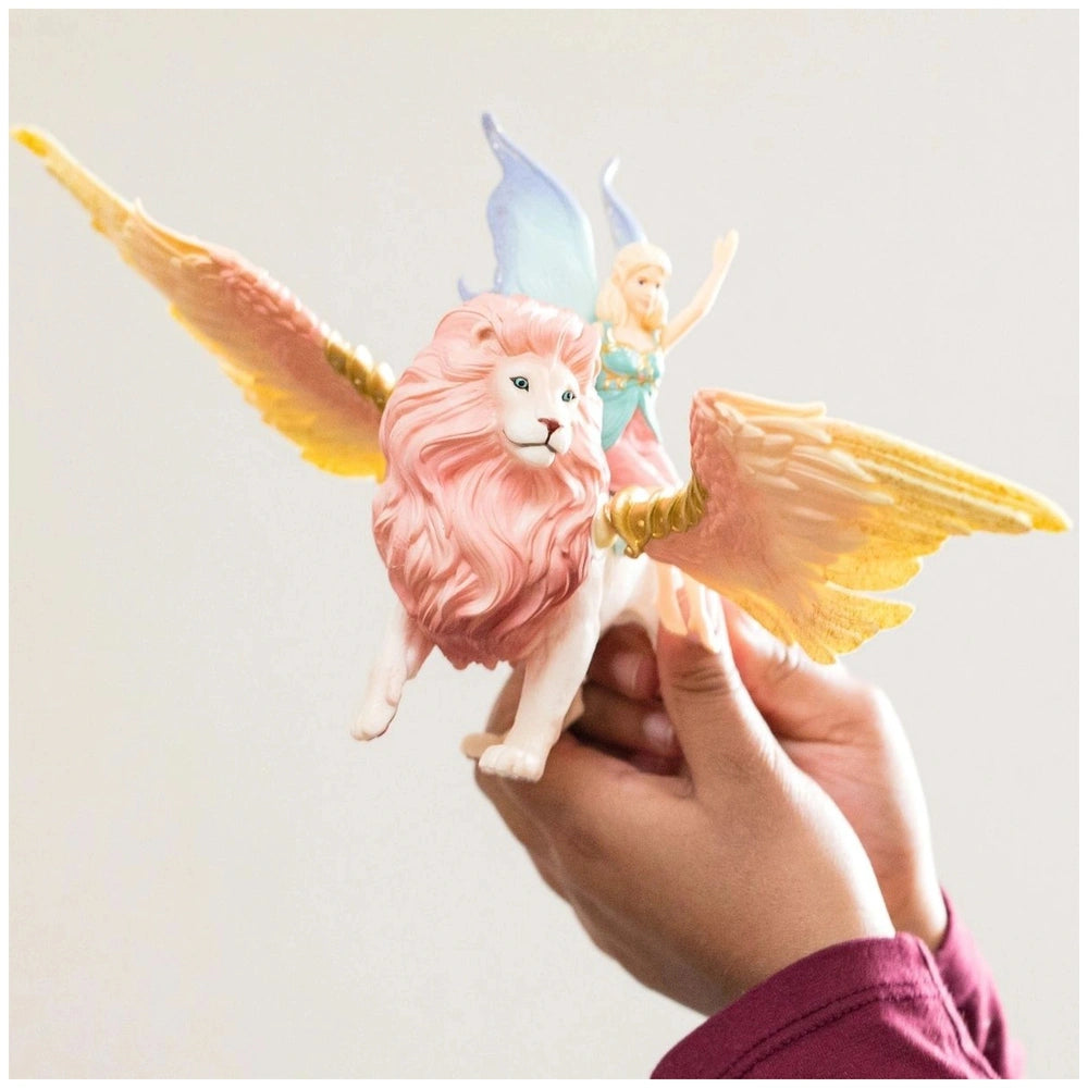 Fairy figurine of a winged lion with a pinkish-red mane and yellow-orange wings held in a hand against a light background.