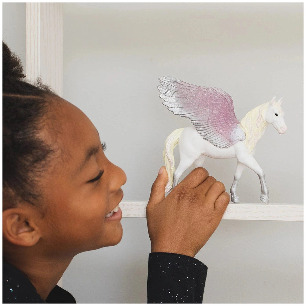 A young girl smiles while holding a white and pink winged horse figurine indoors against a white wall.