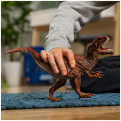 Allosaurus toy dinosaur in brown with a red tail held by a person's hand on a blue rug over a wooden floor.