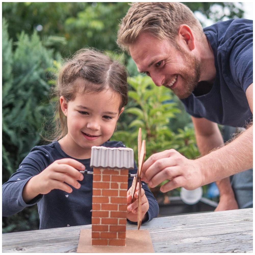 Teifoc windmill arts and crafts scene with a man and girl cutting white paper on a wooden table beside a red brick structure.