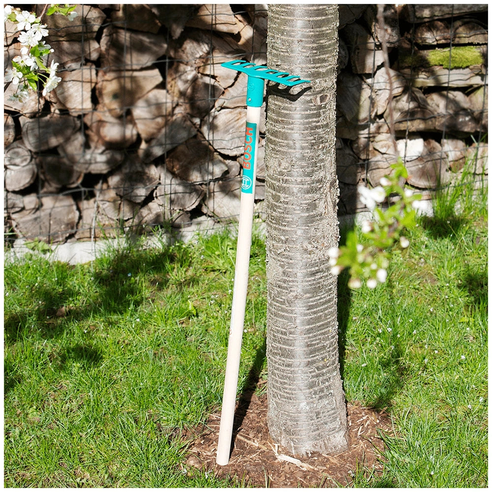 Theo Klein Bosch Garden rake with a white handle and blue grip, placed on green grass near a brown tree trunk and fence.