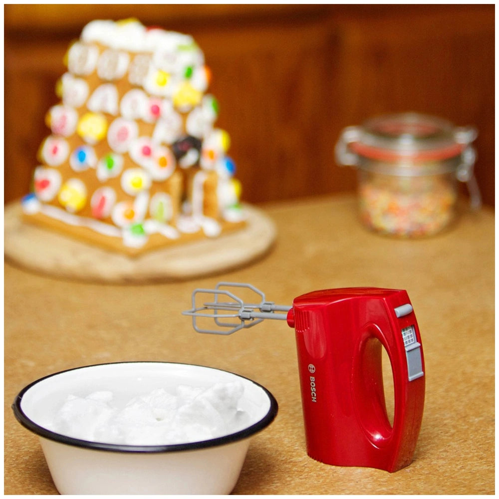 Theo Klein Bosch hand mixer with a red whisk and white bowl on a countertop, featuring a gingerbread house in the background.