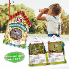 A young child in a white shirt and blue overalls observes a bird through a red telescope in a green outdoor setting.